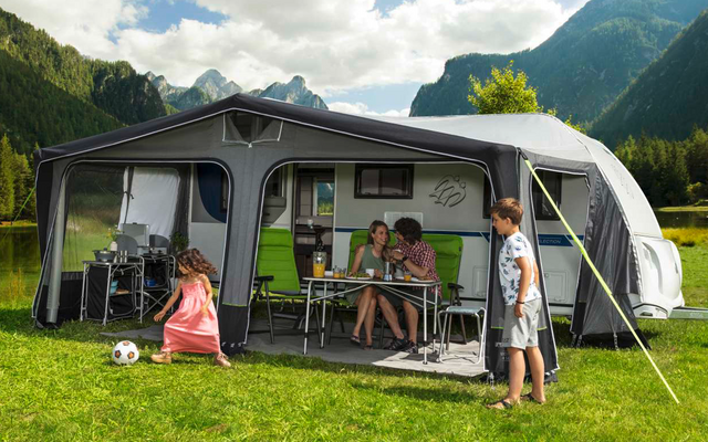 Family in front of a caravan with an attached awning on a green meadow in the mountains