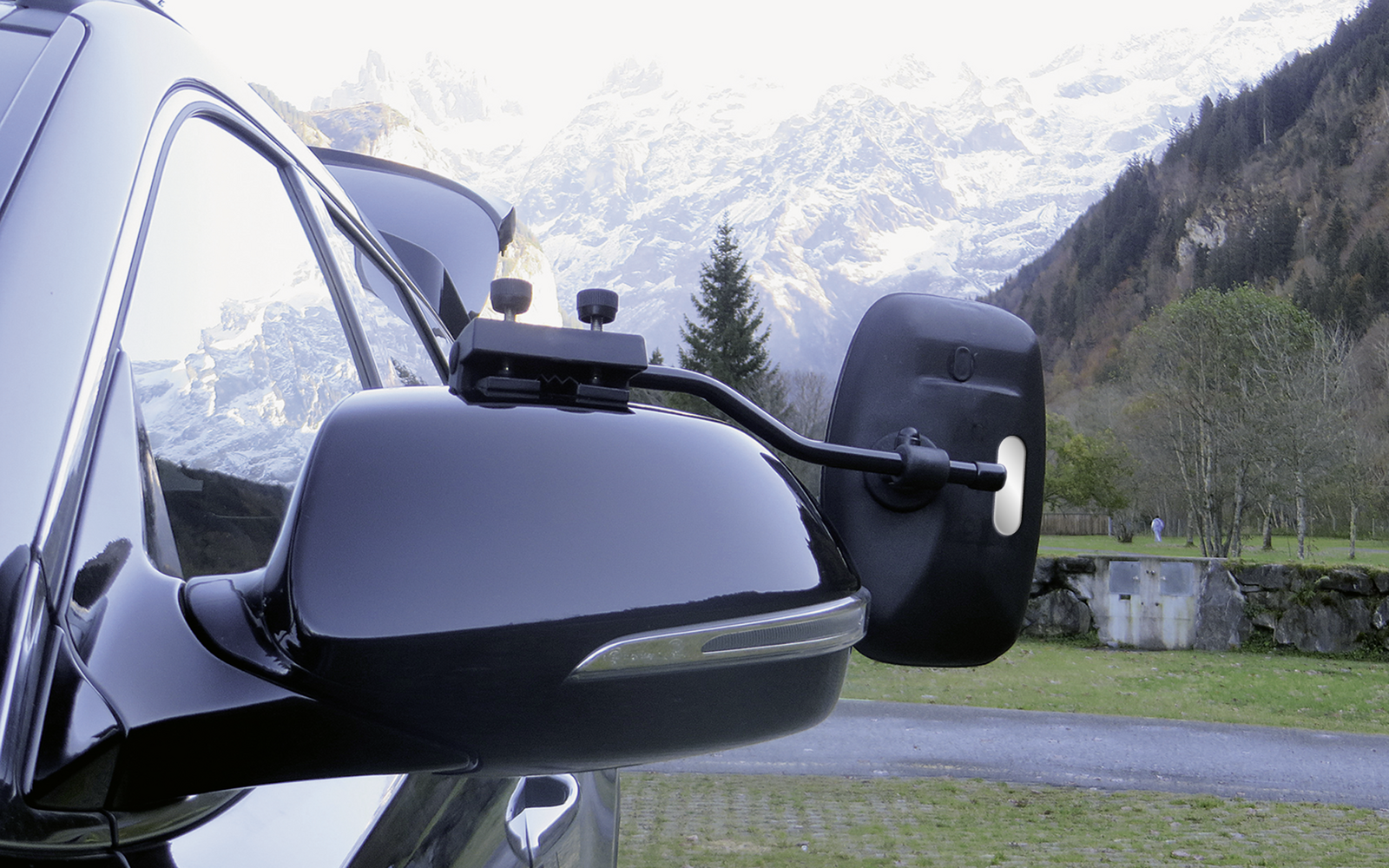 Seitenspiegel eines schwarzen Autos mit montierter Kamera, im Hintergrund Berge und Bäume