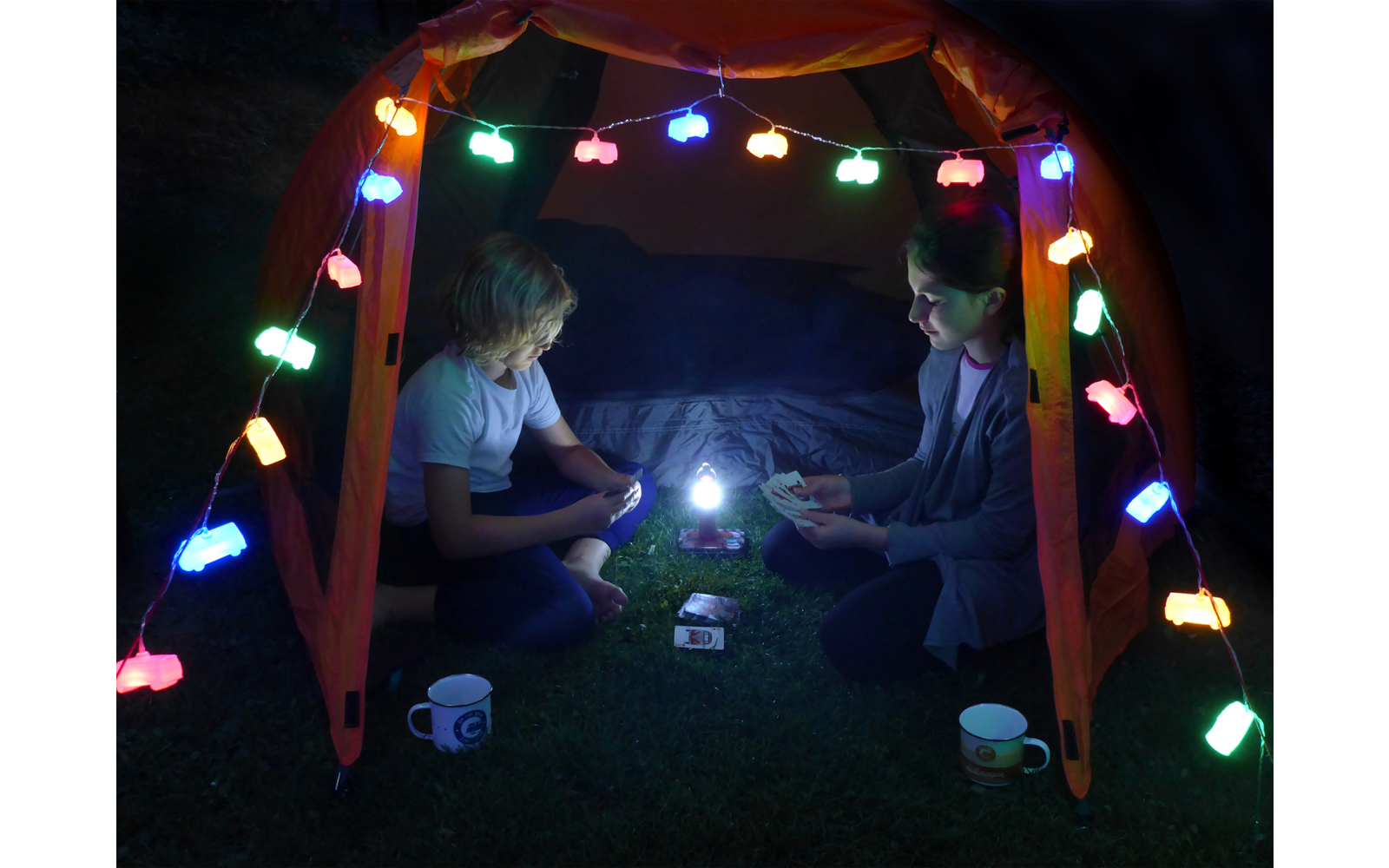 Zwei Kinder sitzen in einem Zelt, das mit bunten Lichterketten beleuchtet ist. Sie halten eine Taschenlampe