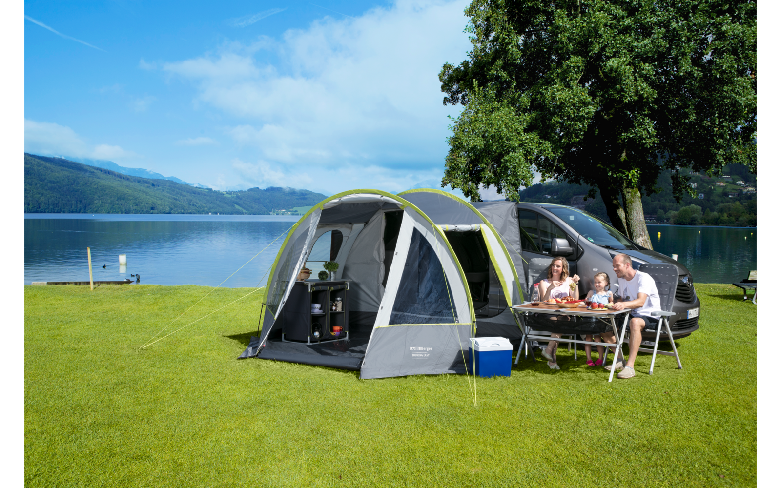 Familie sitzt an einem Campingtisch neben einem Zelt und einem Wohnmobil auf einer Wiese mit Blick auf einen See