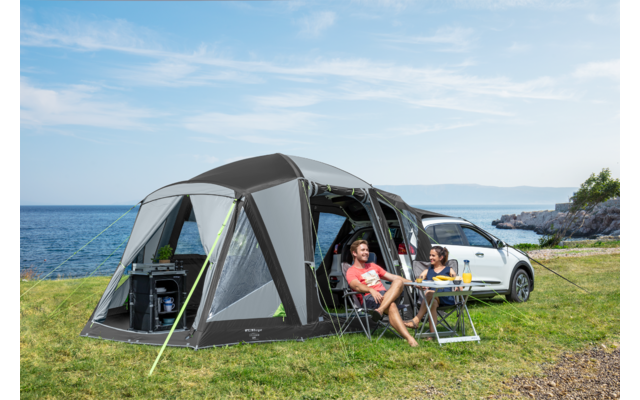 Zwei Personen sitzen vor einem Wohnmobil, das mit einem Zeltvorbau auf einer Wiese neben dem Meer steht