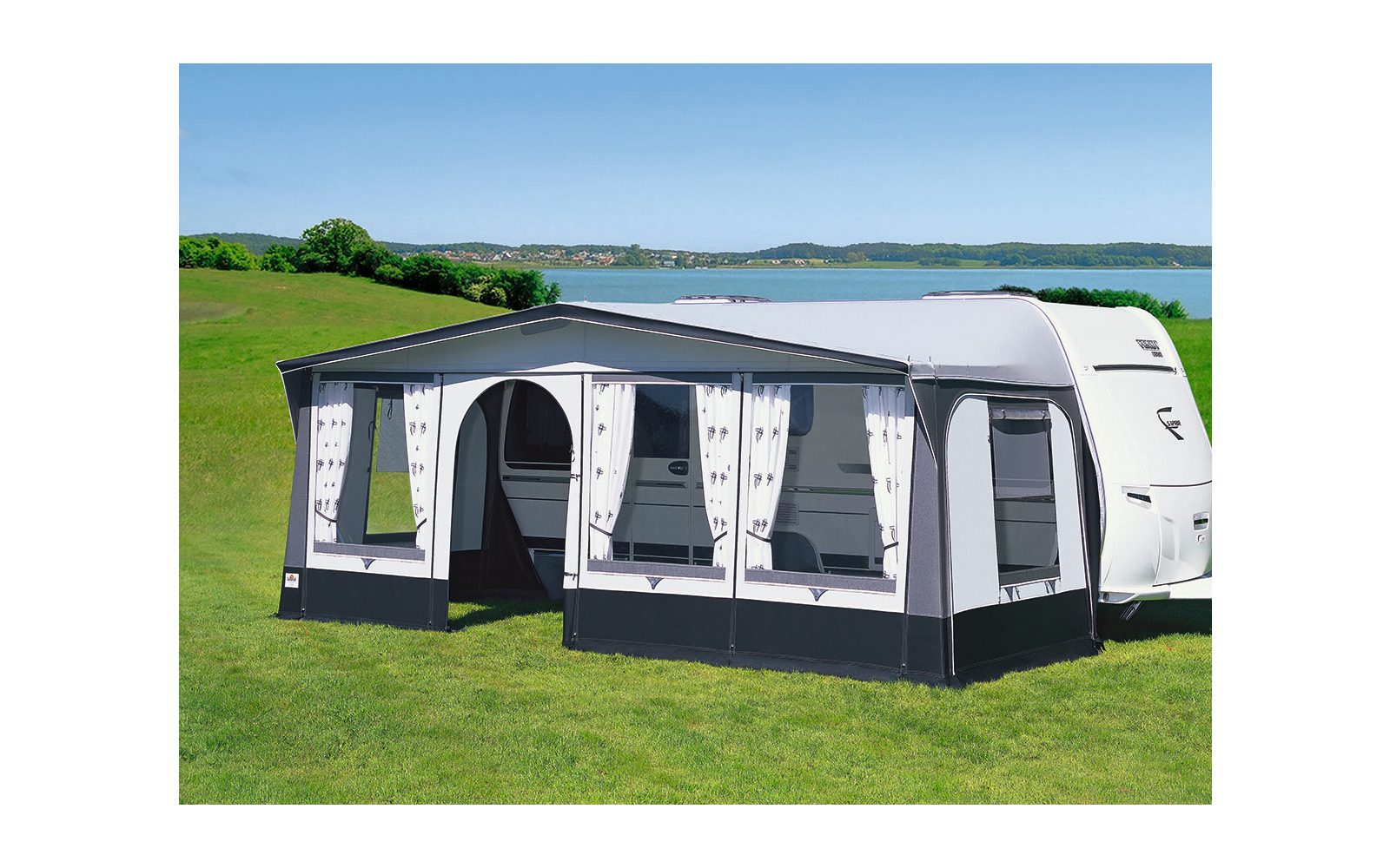 Wohnwagen mit angebautem heller Vorzelt auf einer grünen Wiese vor einem See und blauem Himmel