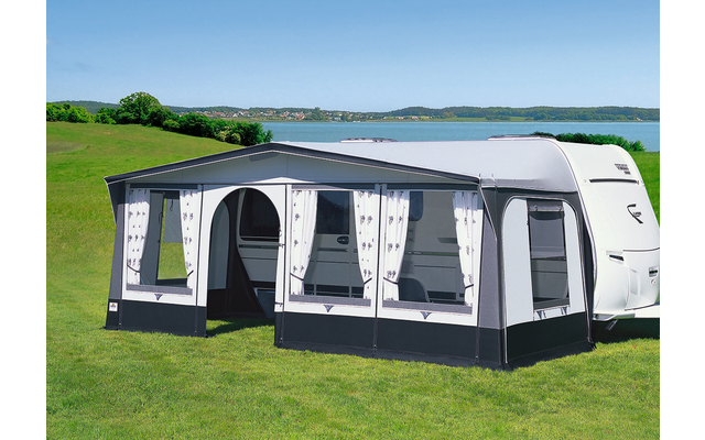 Wohnwagen mit angebautem heller Vorzelt auf einer grünen Wiese vor einem See und blauem Himmel