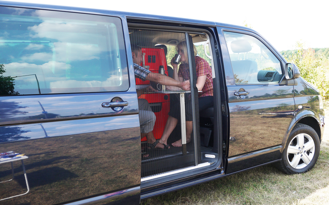 Schwarzer Van mit offener Schiebetür auf einer Wiese, darin zwei sitzende Personen