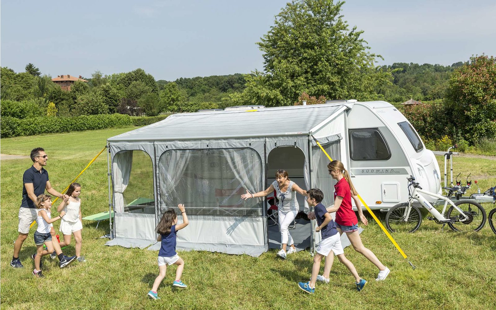 Familie mit Kindern spielt vor einem weißen Wohnwagen auf einer grünen Wiese
