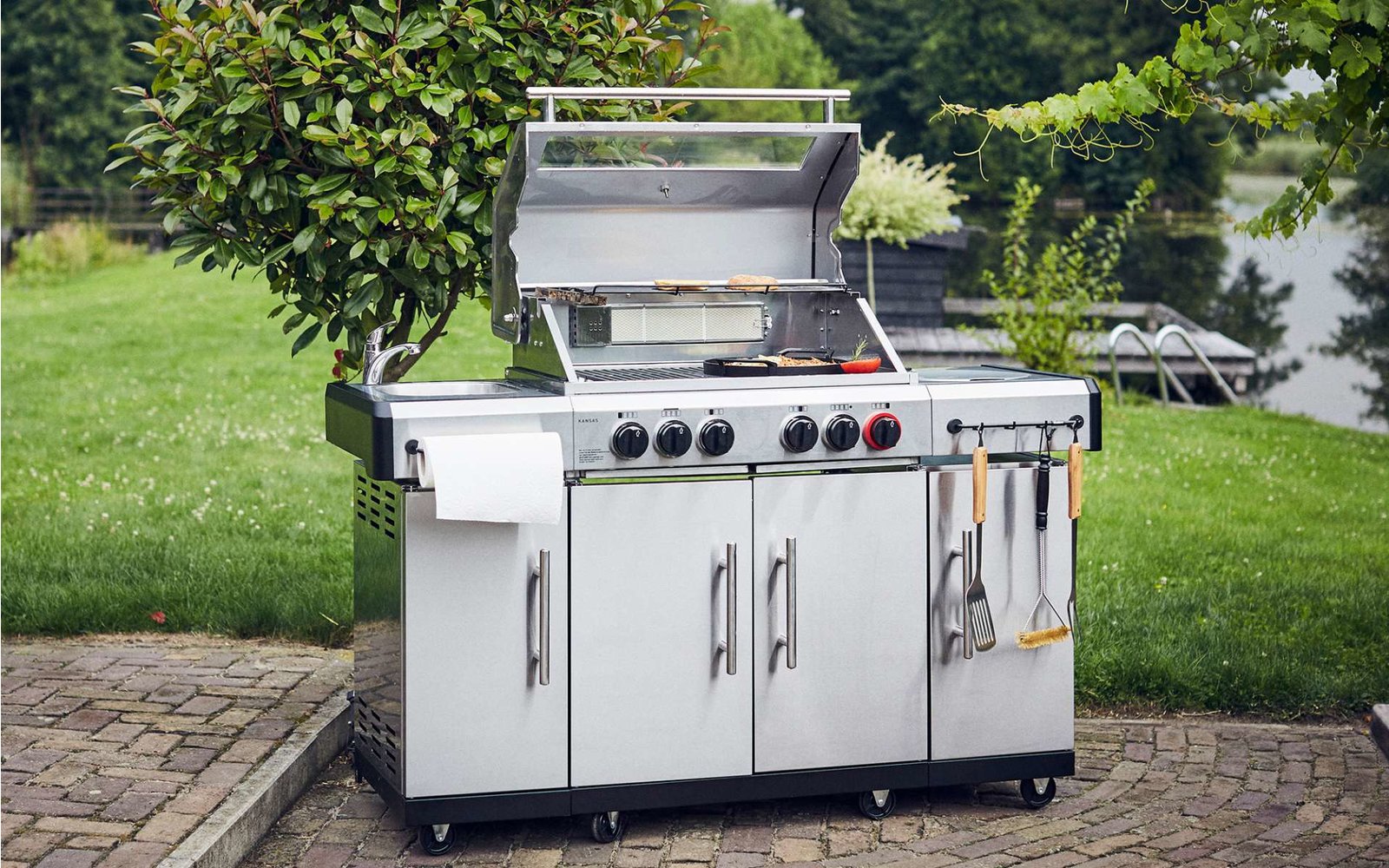 Ein moderner Gasgrill mit vier Brennern und mehreren Schränken steht auf einer gepflasterten Fläche neben einem Rasen