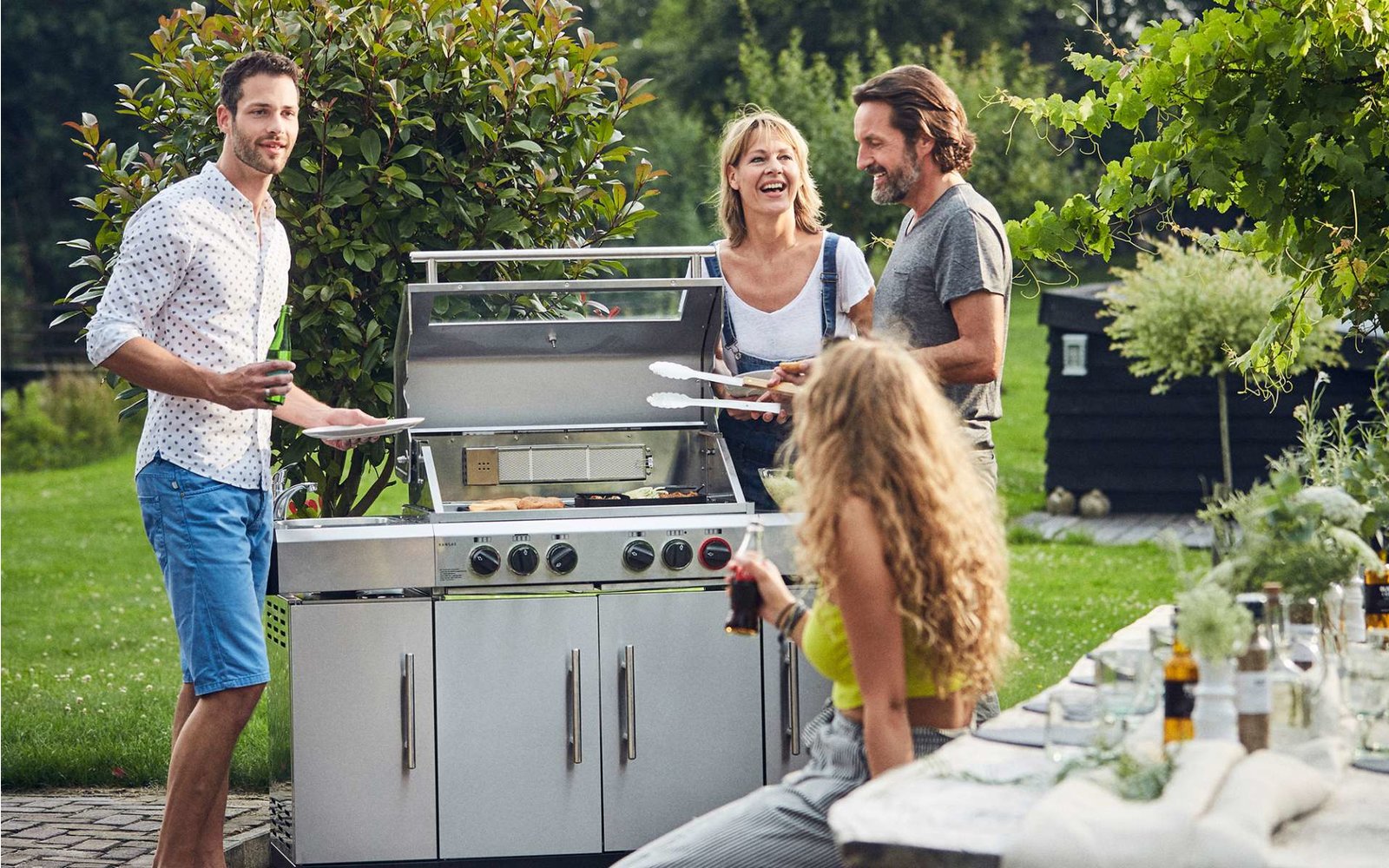 Drei Personen stehen um einen Grill im Garten, zwei sind am Grill, eine kniet daneben
