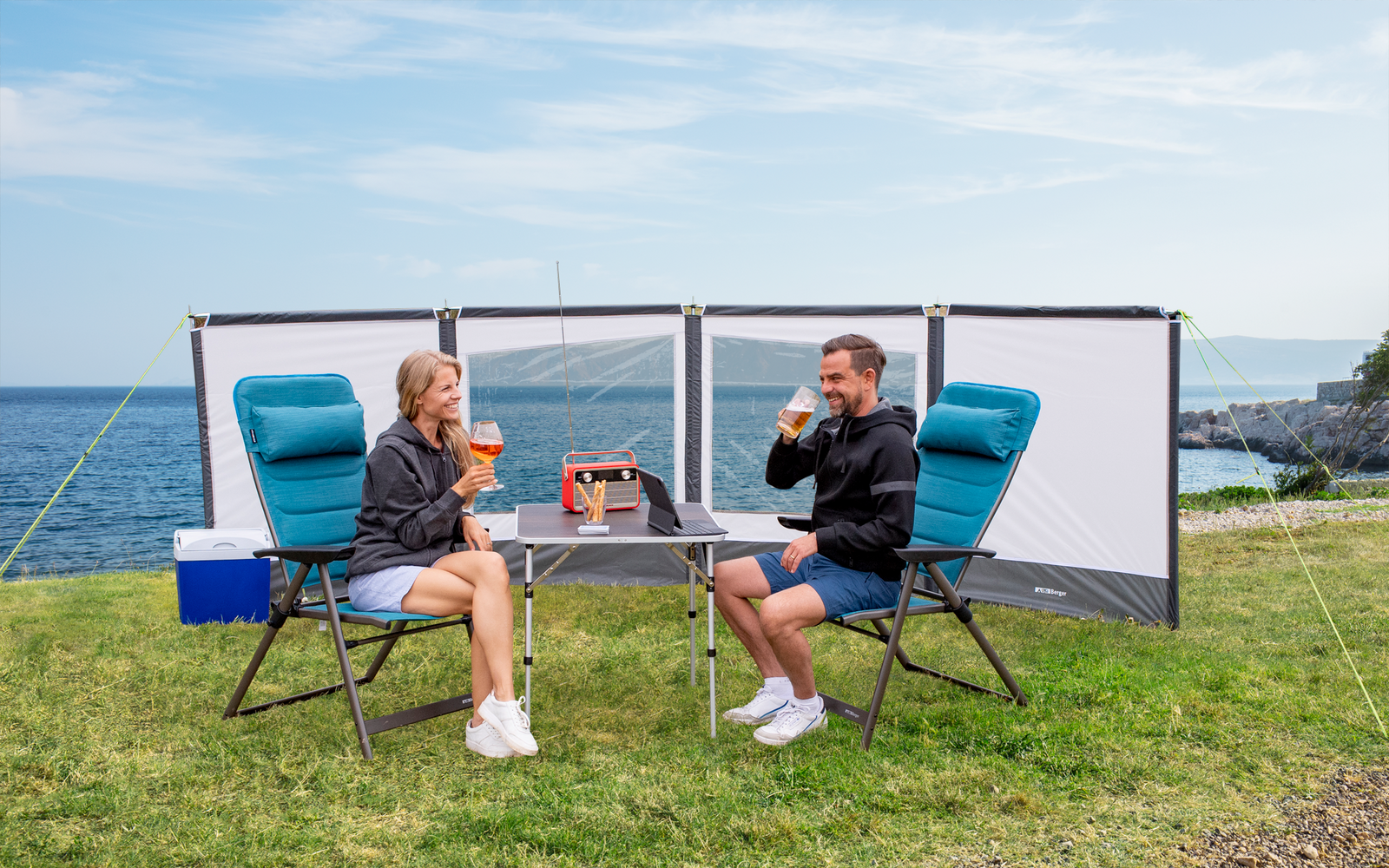 Two people sitting at a camping table with folding chairs on a grassy area by the sea, with a windbreak set up in the background