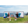 Two people sitting at a camping table with folding chairs on a grassy area by the sea, with a windbreak set up in the background