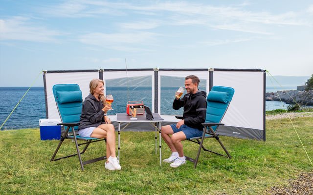 Two people sitting at a camping table with folding chairs on a grassy area by the sea, with a windbreak set up in the background