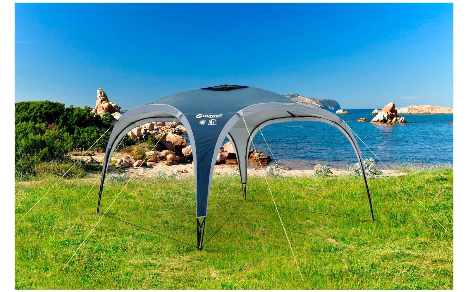 Graues Zelt auf grüner Wiese mit klarem blauen Himmel und Felsen im Hintergrund am Meer