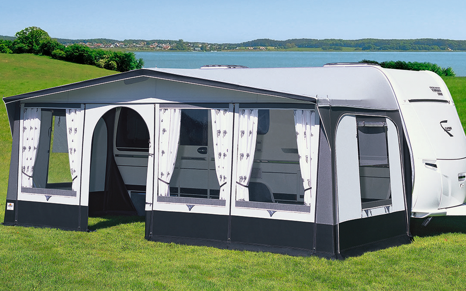 Caravana con toldo claro adjunto en un prado verde frente a un lago y cielo azul