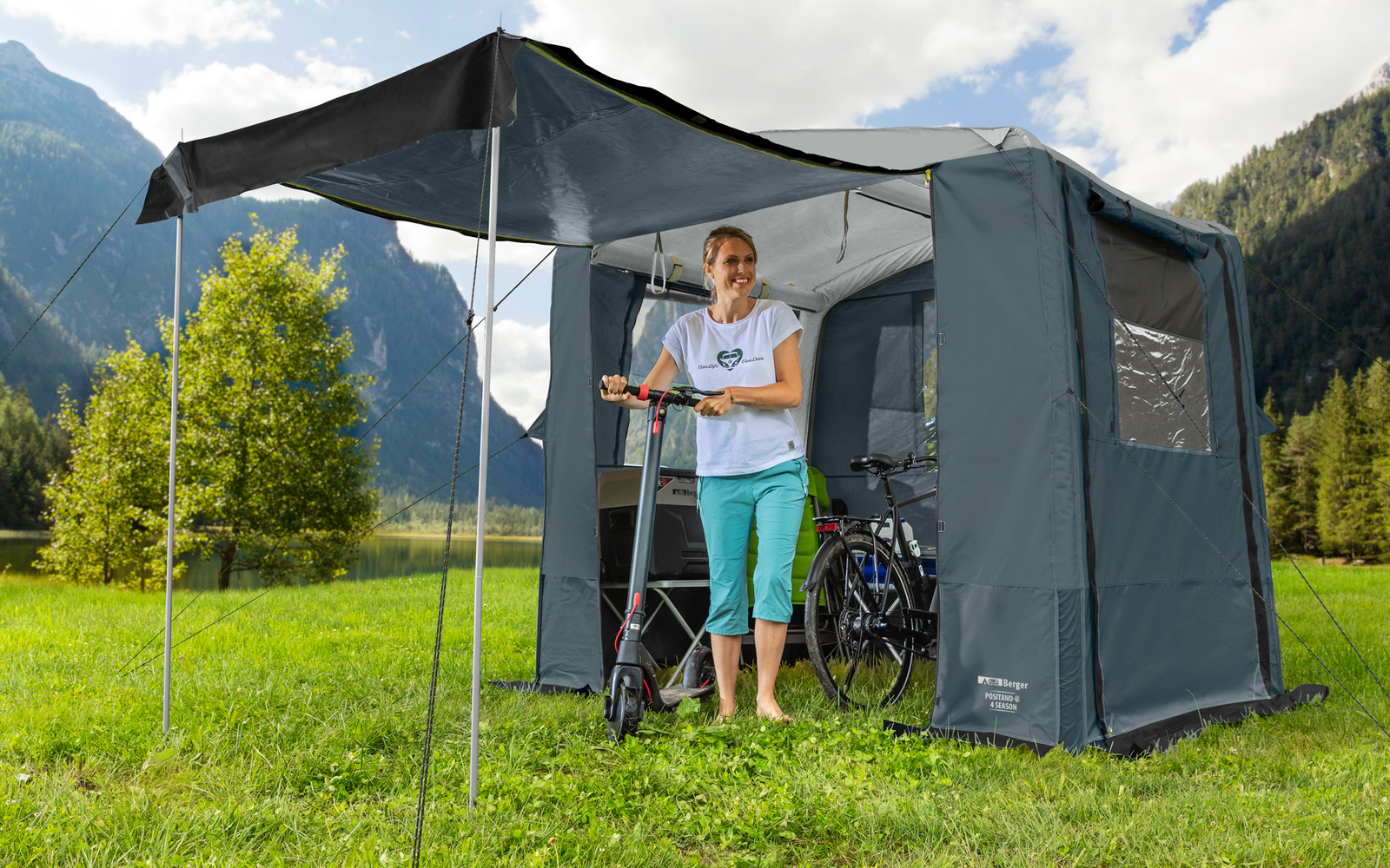 Person steht vor einem grauen Campingzelt auf einer grünen Wiese in den Bergen