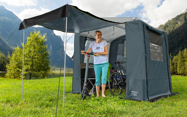 Person steht vor einem grauen Campingzelt auf einer grünen Wiese in den Bergen