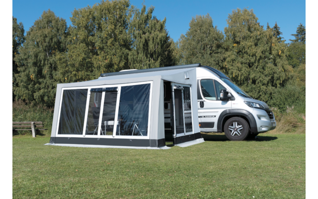 Weißes Wohnmobil mit angebautem, schwarzem Vorzelt auf einer grünen Wiese vor Nadelbäumen