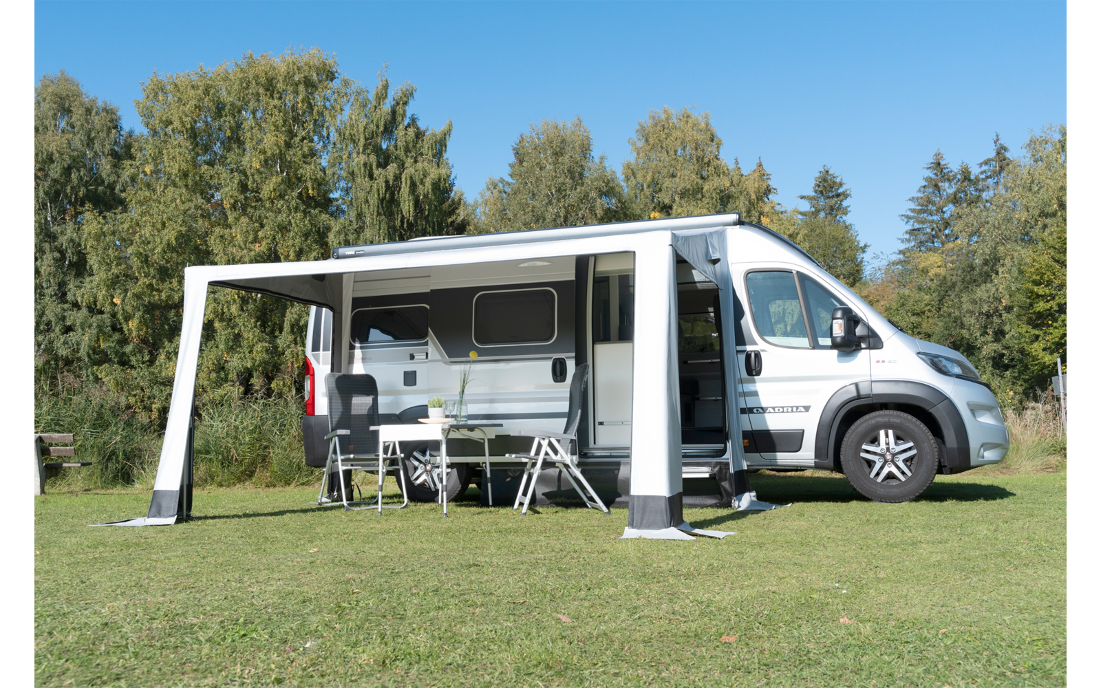 Weißes Wohnmobil mit ausgefahrener Markise und zwei Stühlen auf einer grünen Wiese vor Bäumen