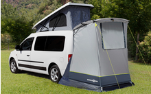 Weißer Van mit aufgestelltem Zeltanhang auf grüner Wiese vor bewaldeten Bergen