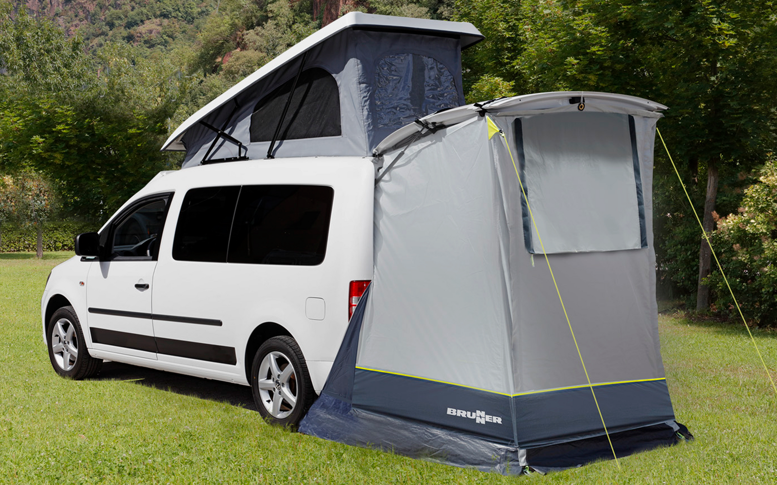 Witte bus met opgezet tentaanhangsel op een groen weiland voor beboste bergen