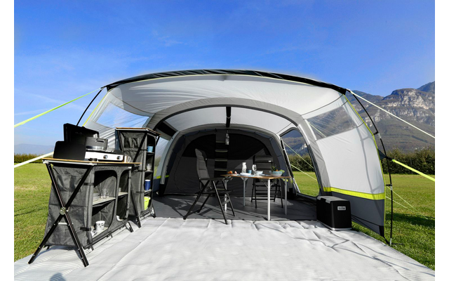 Ein offenes Campingzelt mit Grill, Tisch und Stühlen auf einer Wiese, im Hintergrund Berge