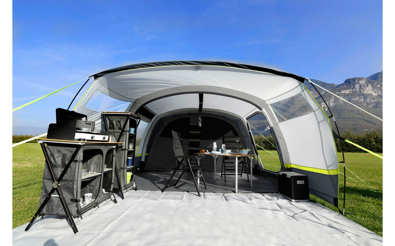 Ein offenes Campingzelt mit Grill, Tisch und Stühlen auf einer Wiese, im Hintergrund Berge