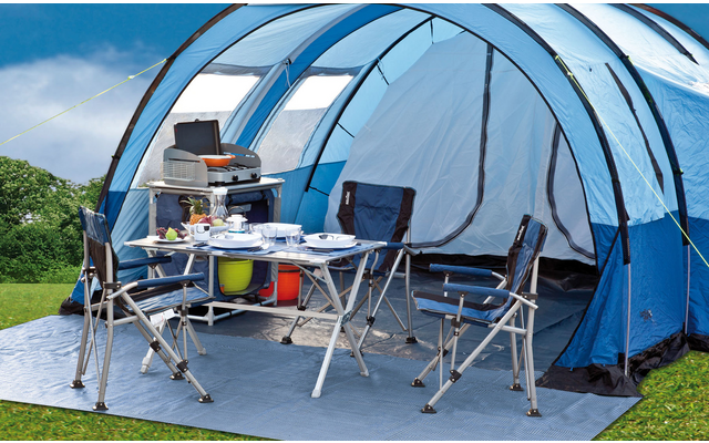 Zwei Campingstühle und ein Campingtisch unter einem blauen Zelt, draußen auf einer Wiese mit Bergen im Hintergrund