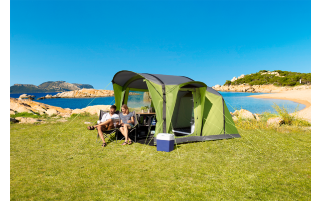 Zelt auf einer Wiese nahe einem Strand, zwei Personen sitzen an einem Tisch unter dem Zelt, blauer Himmel, Küste im Hintergrund
