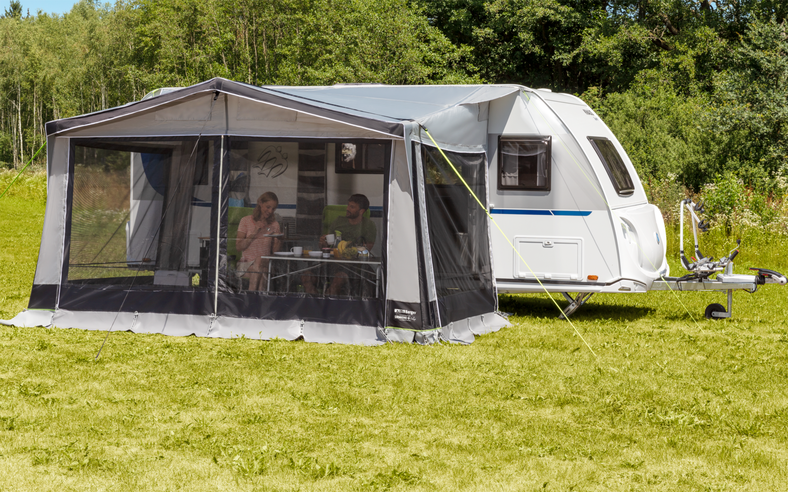 Wohnwagen mit angebautem Zelt auf einer Wiese vor Bäumen