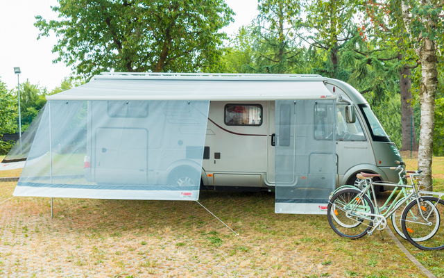 Autocaravana gris estacionada en césped con toldo extendido, dos bicicletas delante