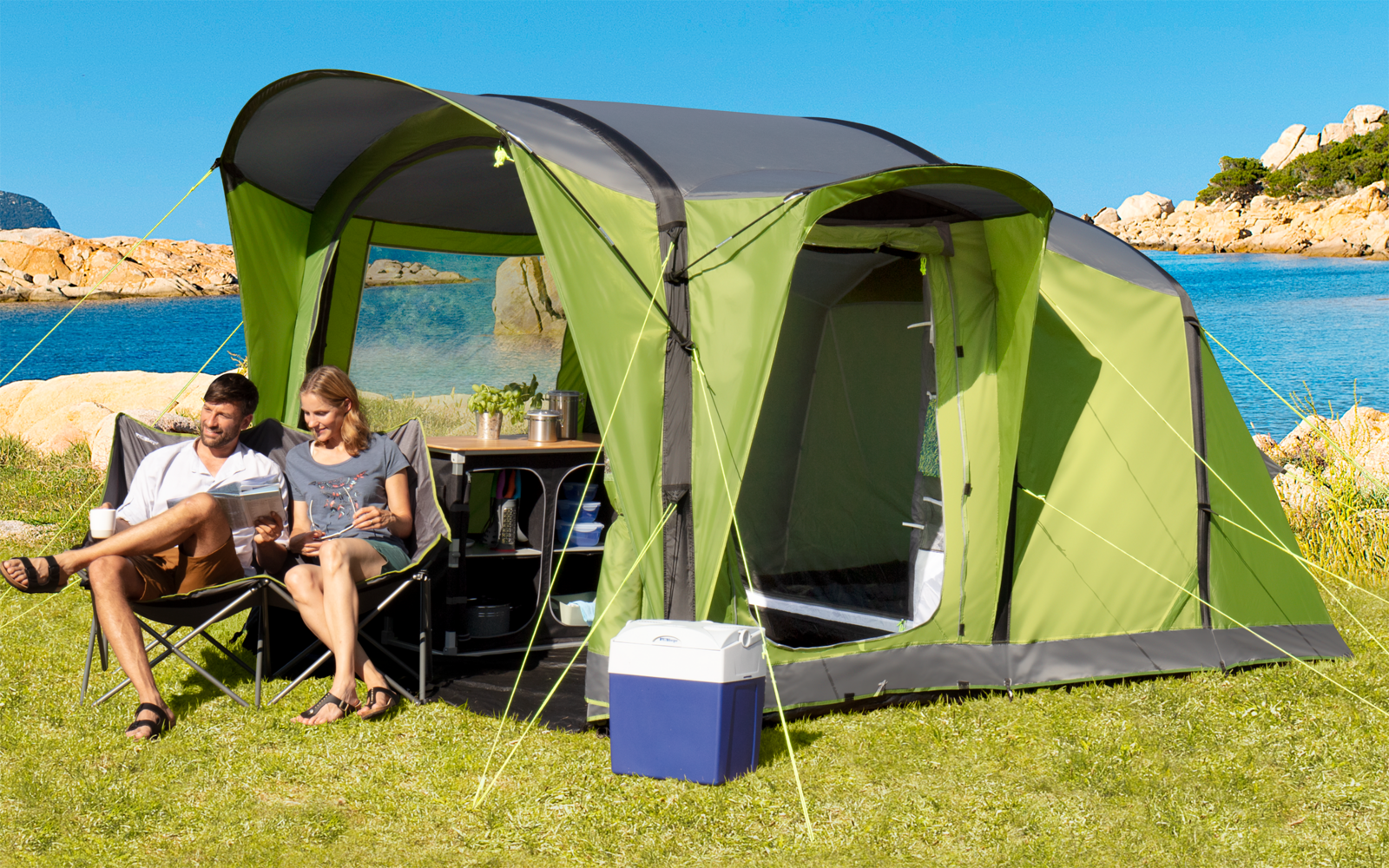 Zelt auf einer Wiese nahe einem Strand, zwei Personen sitzen an einem Tisch unter dem Zelt, blauer Himmel, Küste im Hintergrund