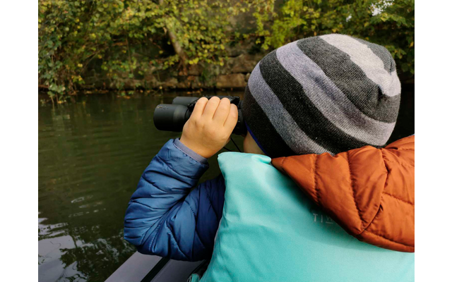 Niño con gorro y prismáticos mirando un lago con árboles al fondo