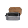 Een open rechthoekige metalen lunchtrommel met een houten deksel op de achtergrond