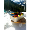 Kom met muesli en fruit, op de achtergrond besneeuwde bergen en dennenbos