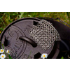 Schwarzer gusseiserner Topf mit Deckel und einem kleinen Edelstahlrost darauf, umgeben von Gras und kleinen weißen Blumen