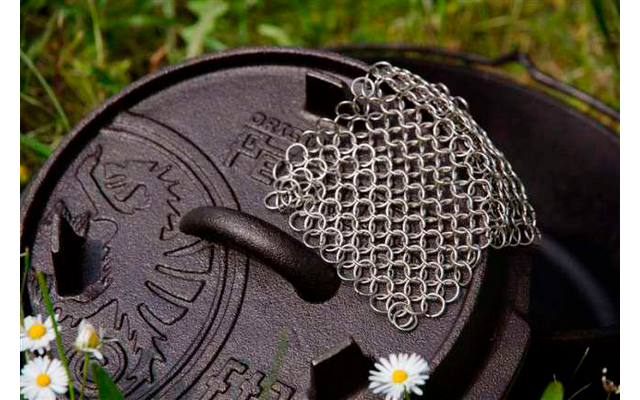 Schwarzer gusseiserner Topf mit Deckel und einem kleinen Edelstahlrost darauf, umgeben von Gras und kleinen weißen Blumen