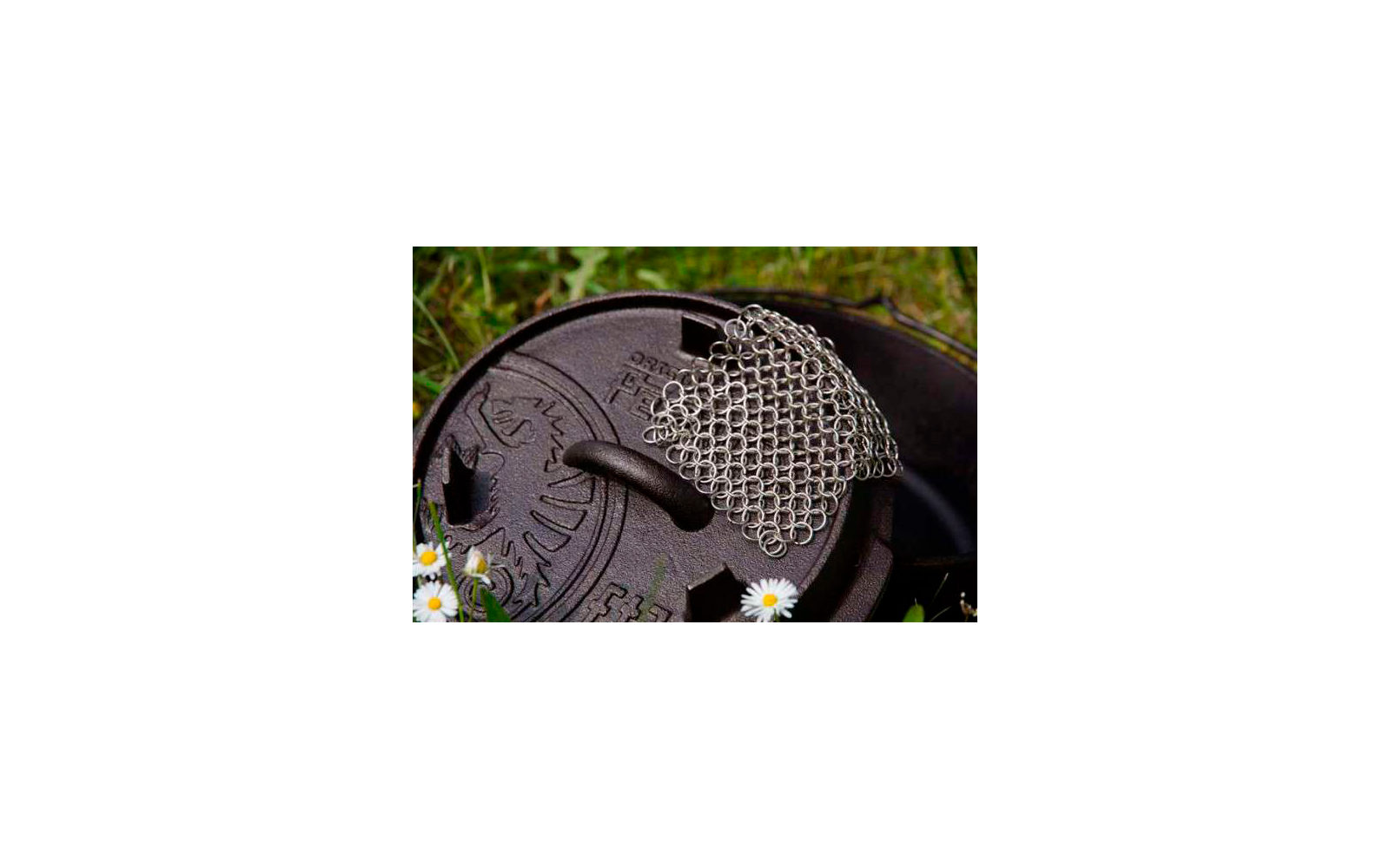 Schwarzer gusseiserner Topf mit Deckel und einem kleinen Edelstahlrost darauf, umgeben von Gras und kleinen weißen Blumen