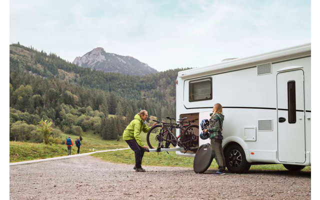 Persoon met rugzak staat naast een witte camper in een bergachtig landschap