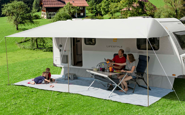 Wohnwagen mit ausgefahrener Markise, zwei Erwachsene sitzen an einem Tisch, ein Kind liegt auf der Wiese