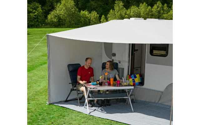 Drei Personen sitzen an einem Tisch unter einem Sonnenschirm vor einem Wohnmobil