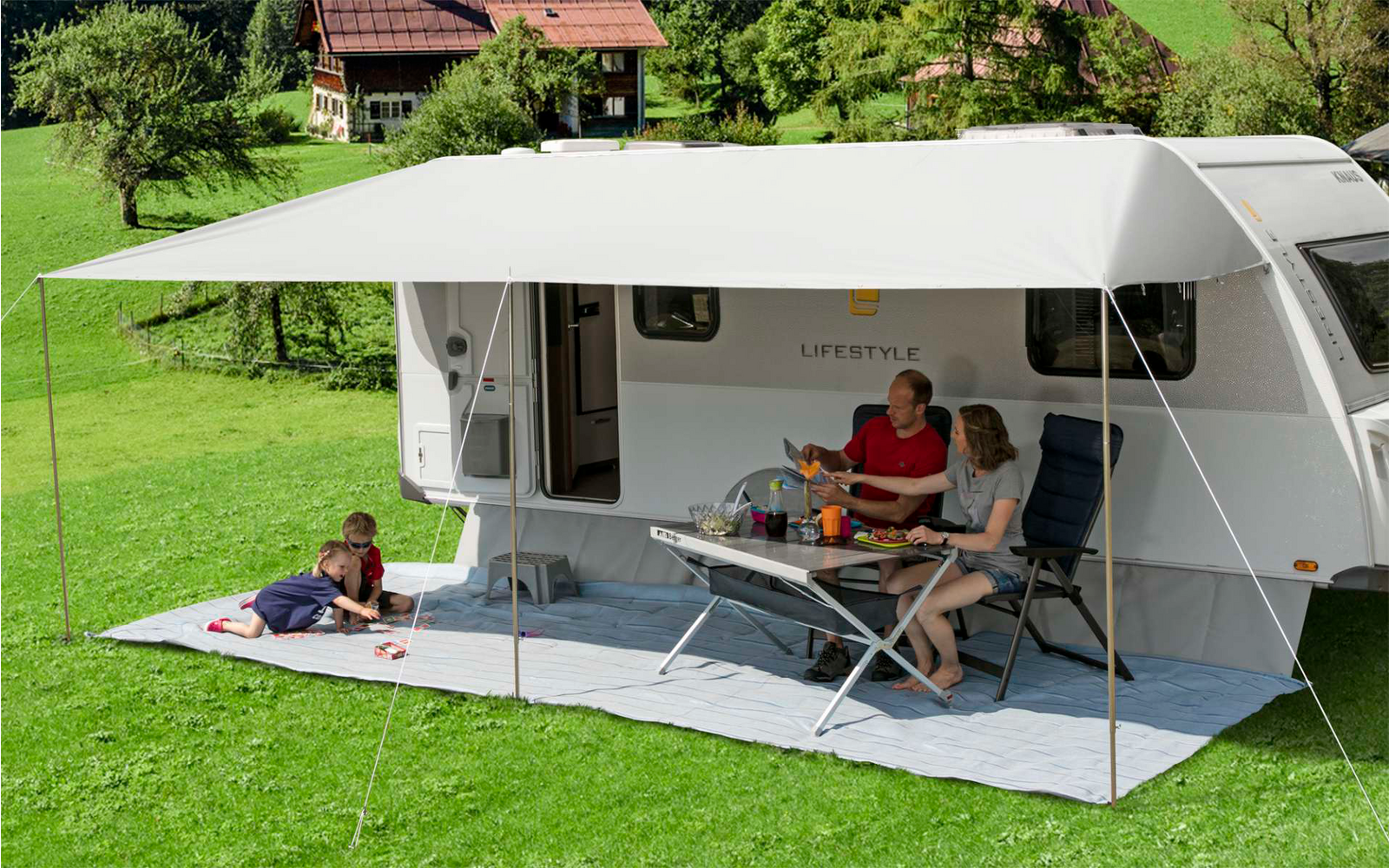 Wohnwagen mit ausgefahrener Markise, zwei Erwachsene sitzen an einem Tisch, ein Kind liegt auf der Wiese