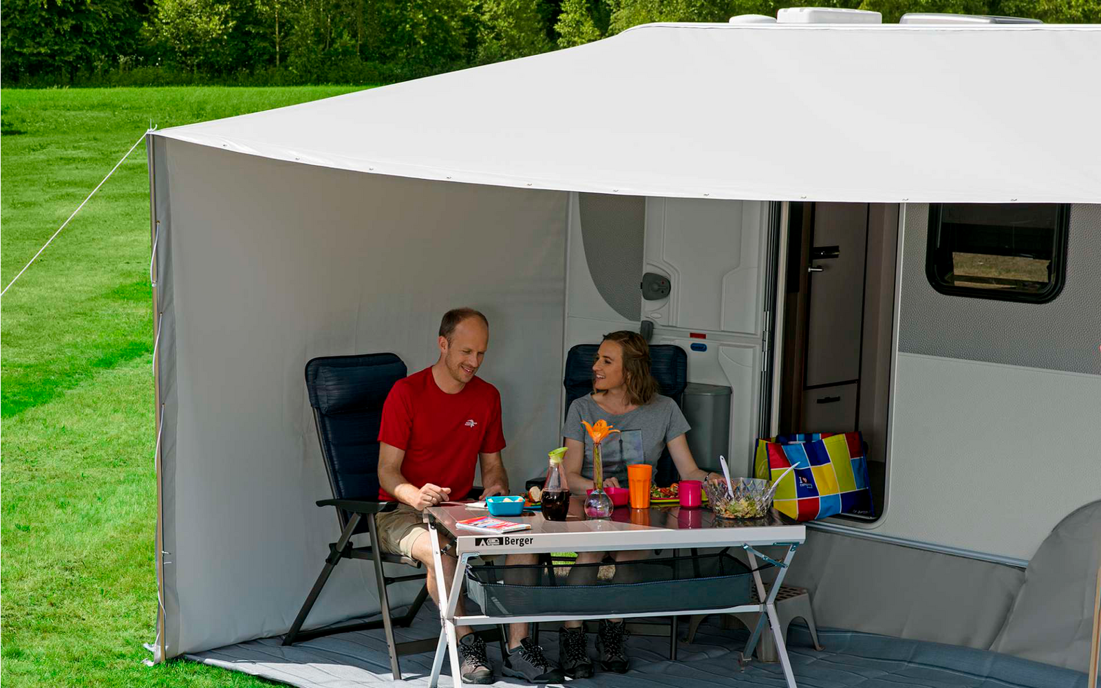 Drei Personen sitzen an einem Tisch unter einem Sonnenschirm vor einem Wohnmobil
