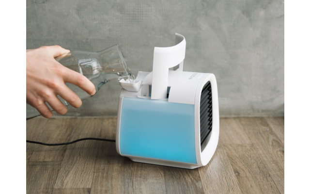 Hand füllt Wasser in kleines elektrisches Gerät mit blauem Wasserbehälter auf Holztisch