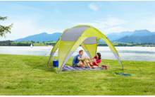 Zwei Personen sitzen auf einer Decke unter einem gelben und grauen Sonnenschutz auf einer Wiese, im Hintergrund Berge und ein See
