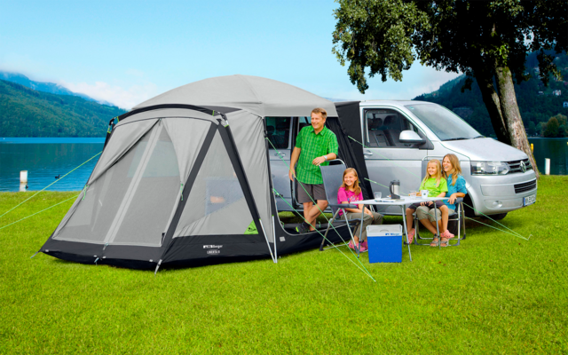 Twee personen zitten aan een kampeertafel voor een grijze tent op gras, met bomen en een blauwe lucht op de achtergrond