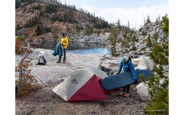 Zwei Personen beim Aufbau eines Zeltes an einem Bergsee mit Felsen und Bäumen im Hintergrund