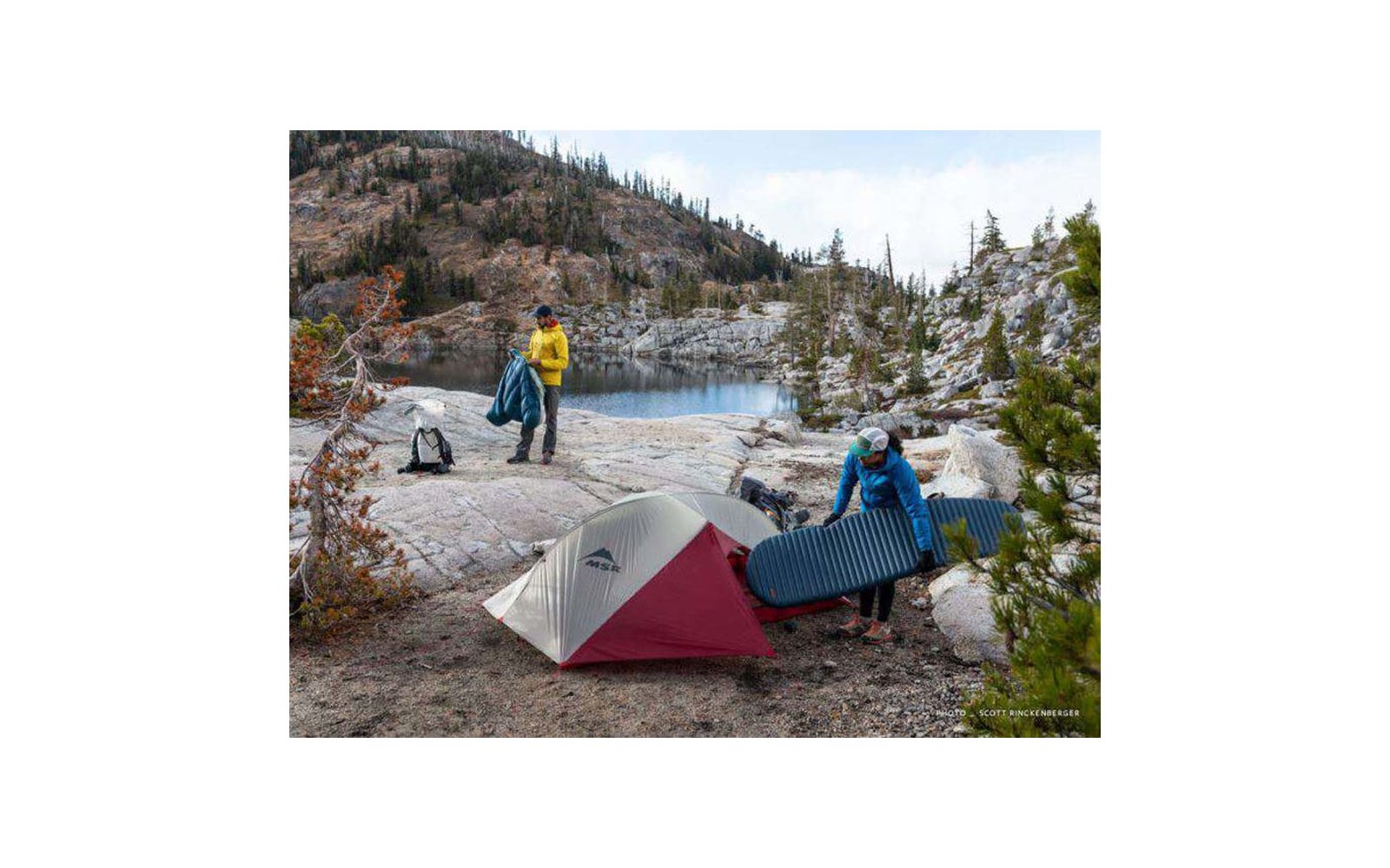 Zwei Personen beim Aufbau eines Zeltes an einem Bergsee mit Felsen und Bäumen im Hintergrund