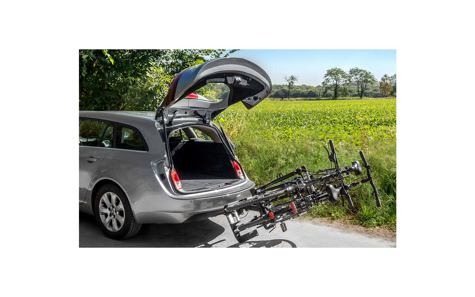 Silberner Kombi mit offenem Kofferraum, Fahrrad am Heck befestigt, grüne Feldlandschaft im Hintergrund