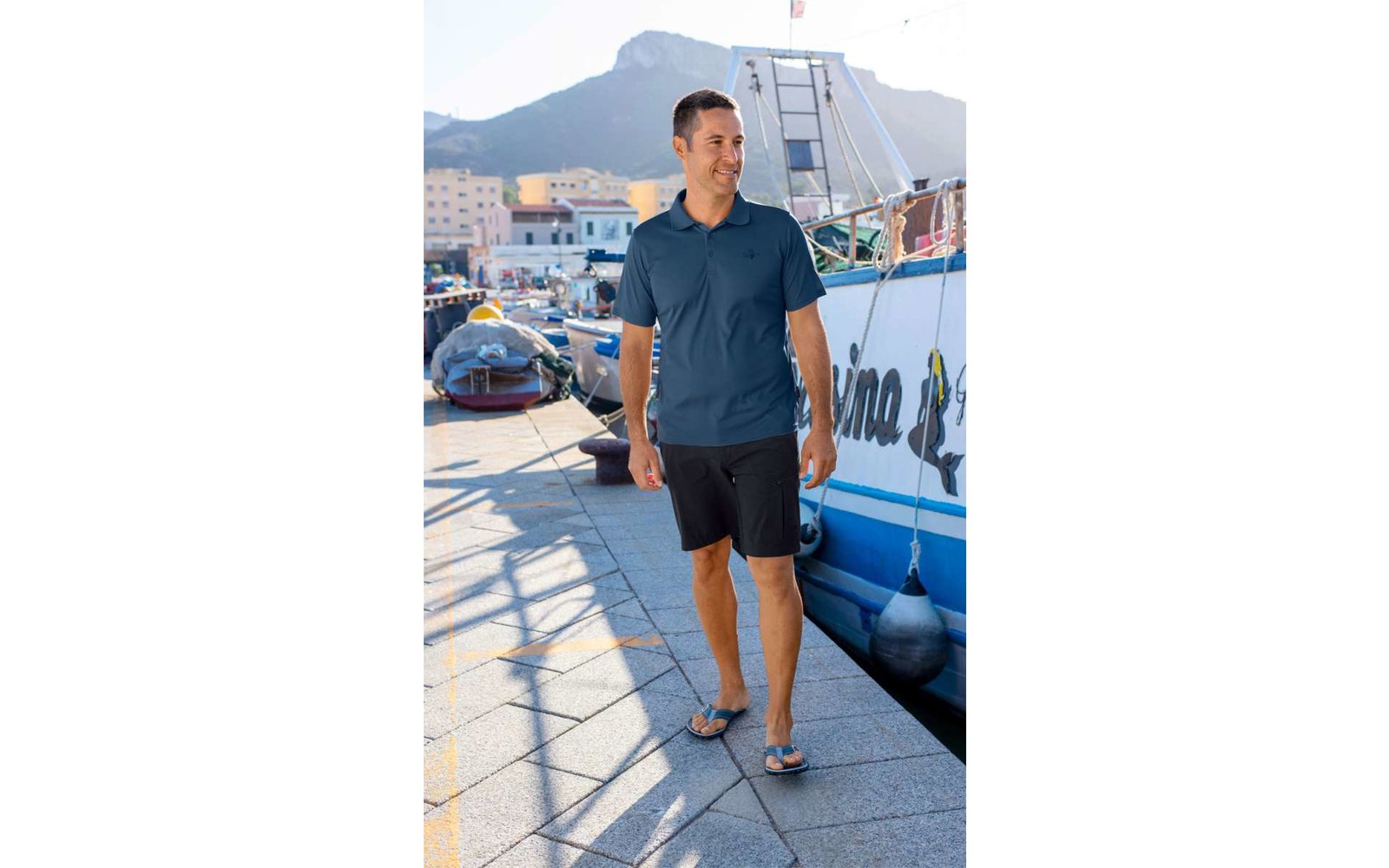 Mann in blauem Hemd und dunkler Hose läuft auf einem gepflasterten Weg entlang von Booten im Hafen
