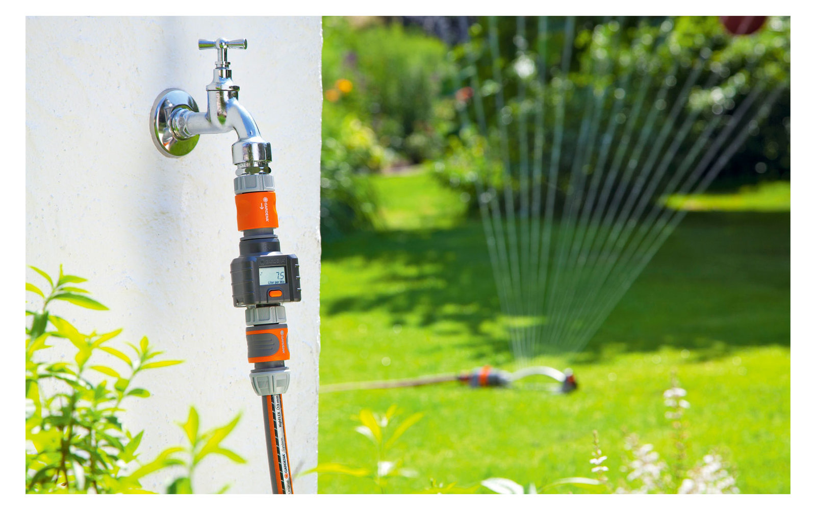 Water faucet with attached garden hose on a white wall, a sprinkler sprays water on a green lawn in the background