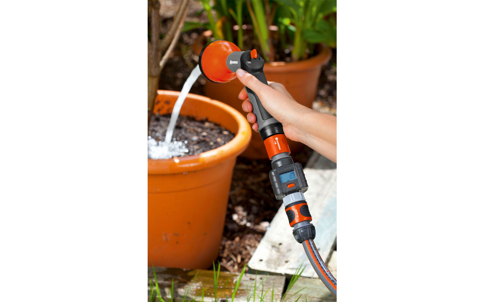 Hand holding garden hose with nozzle watering a plant in a terracotta pot