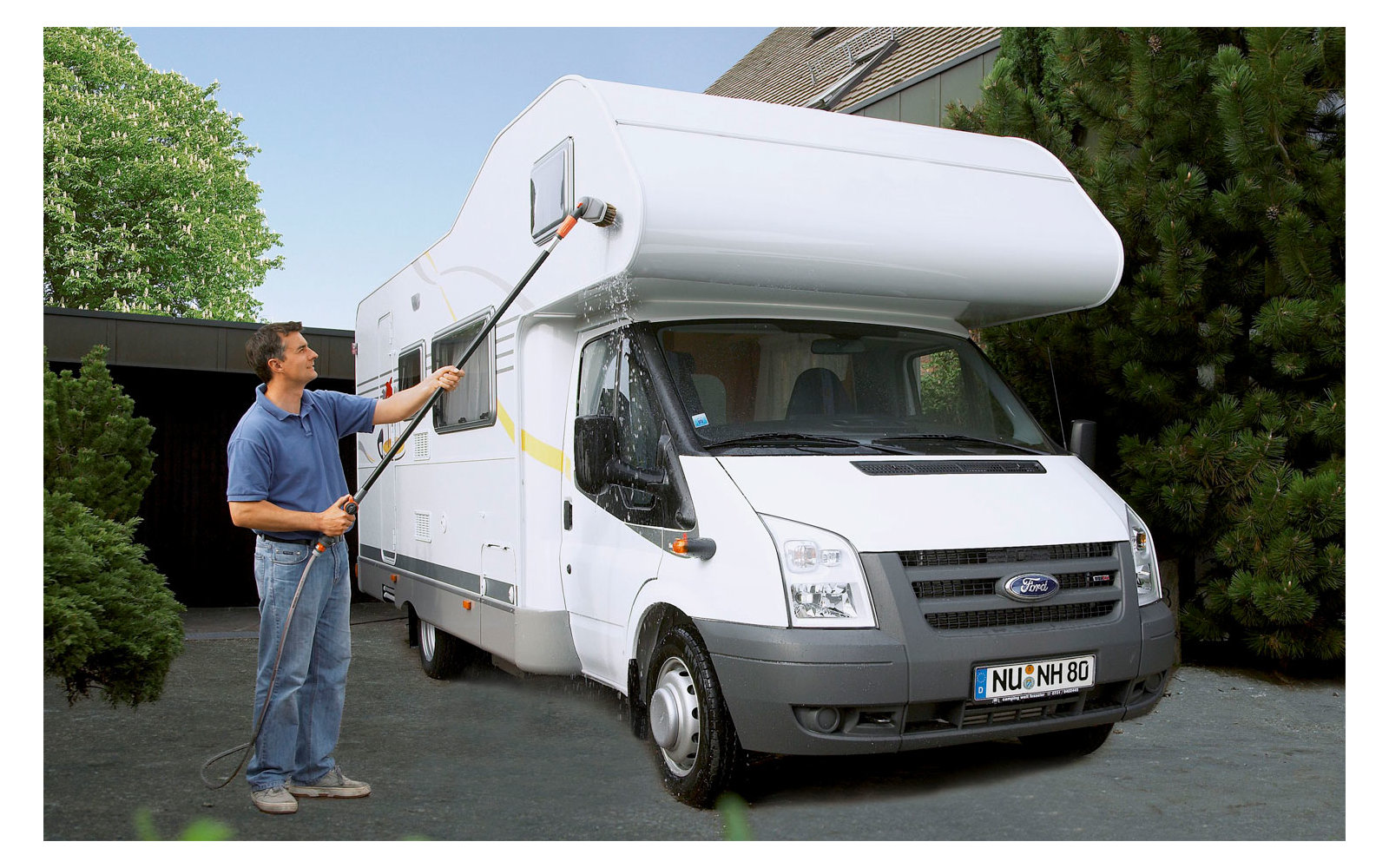 Man cleans the roof of a white camper van with a long telescopic pole in front of a house